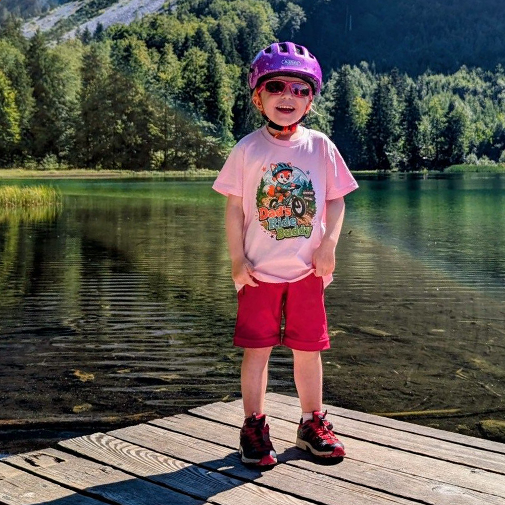 Kind mit Pedal Passion Dad's Ride Buddy T-Shirt, Fahrradhelm und Sonnenbrille auf einem Holzsteg an einem See.